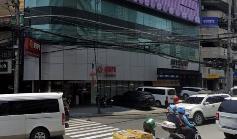External view of the BPI Binondo Plaza Lorenzo Ruiz branch located in a multi-story glass building, featuring prominent red BPI signage above the entrance and on a street-side pole, with city traffic including vans and motorcycles in the foreground