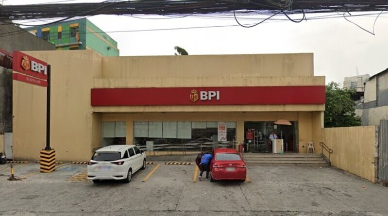 An external view of the BPI Bustillos branch building, featuring its signature red storefront signage with the gold crown logo. A white car and a red car are parked in the front lot, and a security guard stands near the main glass entrance