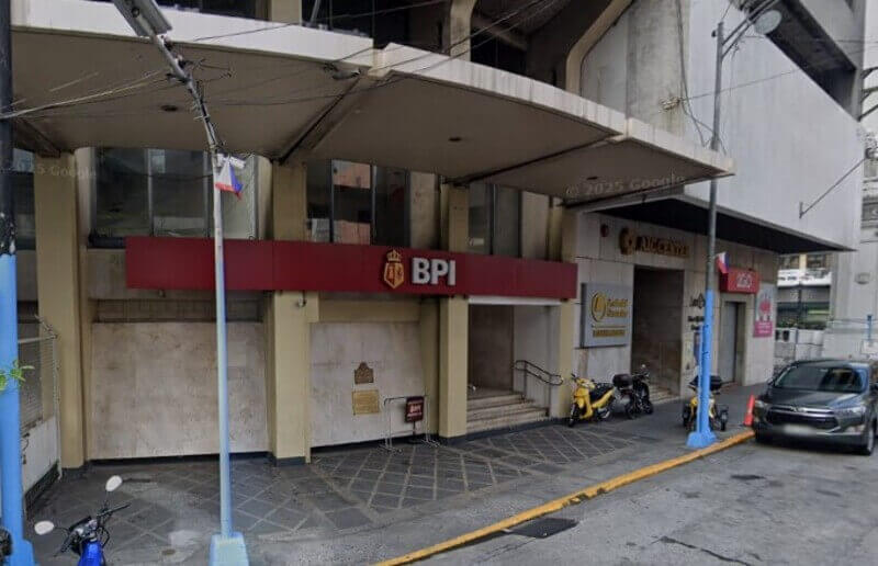 Street-level view of a BPI (Bank of the Philippine Islands) branch entrance featuring a prominent red sign with the gold BPI logo. The building has a modern concrete overhang, marble-textured walls with a historical marker, and is situated next to the AIC Center with parked motorcycles and a car on the street in front