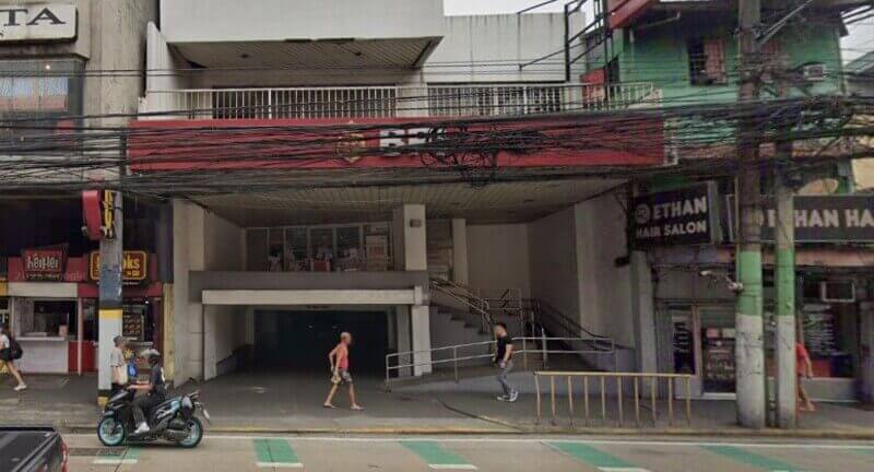 Exterior view of the BPI Legarda branch, featuring a red sign and an entrance with a ramp and stairs, alongside neighboring storefronts and overhead power lines