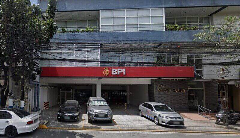 Street view of a BPI branch located on the ground floor of Naess House, featuring a prominent red storefront sign and several cars parked in front