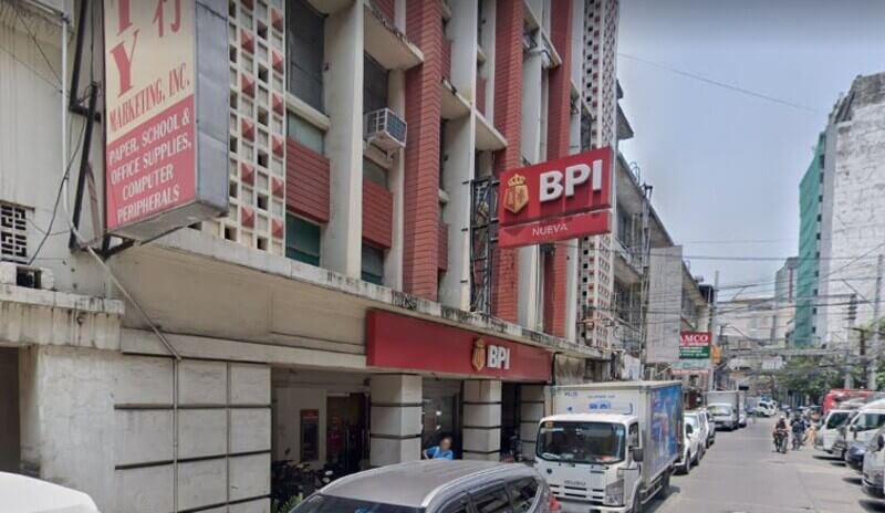 Street view of a red BPI bank branch with a "Nueva" sign on a busy city street in the Philippines, featuring parked cars, a delivery truck, and nearby commercial buildings