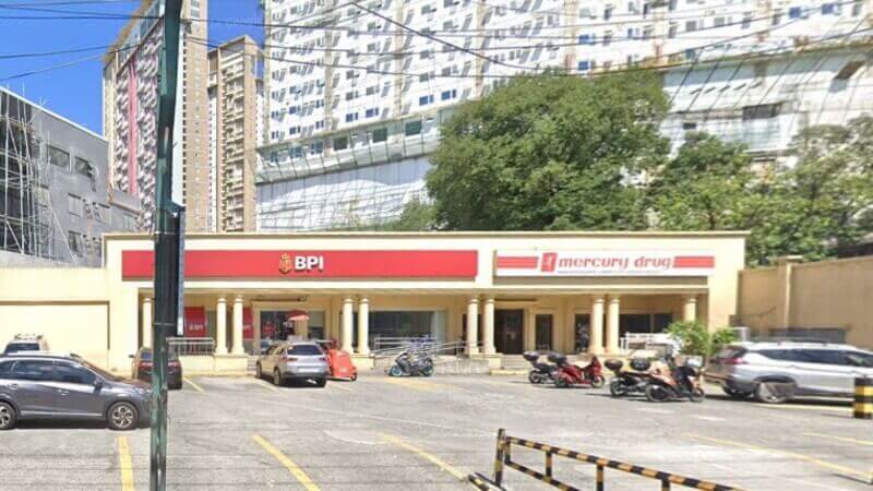 A BPI branch and a Mercury Drug store sharing a single-story commercial building. The building features light yellow walls, classic white pillars, and a spacious outdoor parking area with several cars and motorcycles in the foreground. Tall modern buildings and green trees are visible in the background under a clear blue sky
