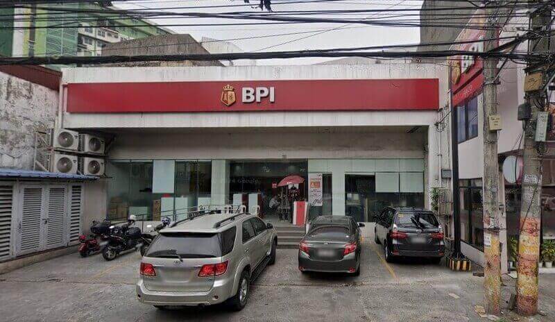 Exterior view of a BPI branch Paco featuring a large red sign with the white BPI logo, a small storefront parking area with several cars and a motorcycle, and various overhead utility lines