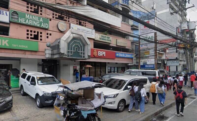 A busy street-level view of the BPI Padre Faura - Taft Avenue branch in Manila, showing the red bank storefront next to an Uncle John’s convenience store, with pedestrians, parked cars, and a tricycle in the foreground