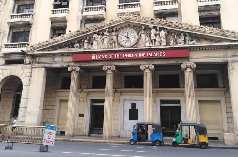 Exterior of the historic BPI Quiapo branch, featuring Neoclassical architecture with fluted Ionic columns, a decorative pediment with sculptures surrounding a central clock, and two colorful e-trikes parked in front