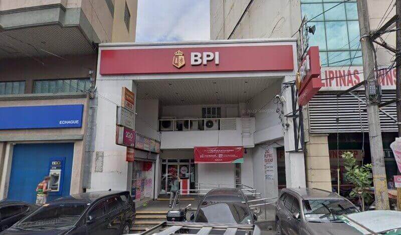 Exterior view of a BPI Quiapo Church branch office featuring a prominent red logo sign, situated on a busy street with parked cars and adjacent commercial buildings
