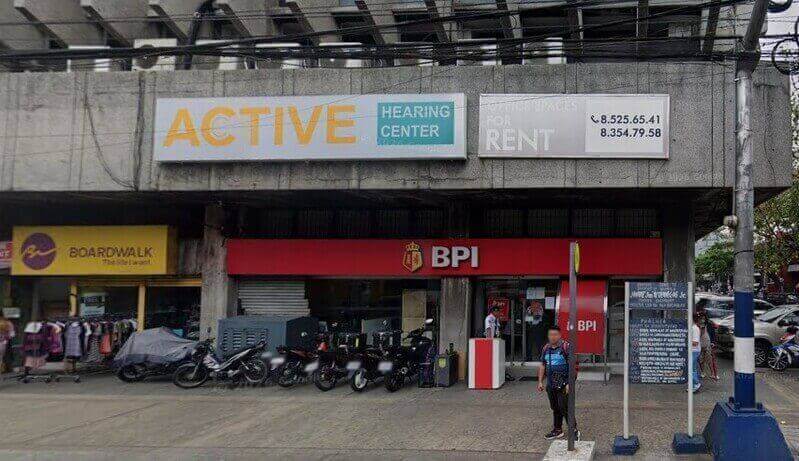 A street-level view of a BPI Remedios branch located on a city sidewalk. To the left is a Boardwalk retail store with a yellow sign, and above the bank are large signs for "Active Hearing Center" and "Office Spaces for Rent." Several motorcycles are parked in front of the building