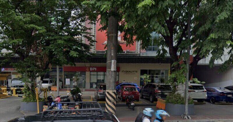 Street view of a BPI branch storefront located on the ground floor of a commercial building, partially obscured by lush green trees and a utility pole. Several cars and motorcycles are parked in front of the entrance, and a "Qualifon" sign is visible next to the red BPI logo