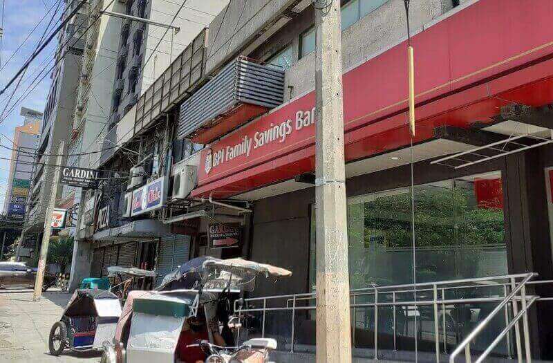 A street-level view of the BPI Family Savings Bank Sampaloc branch, featuring the building's red exterior signage, glass facade, and a metal accessibility railing, with tricycles parked along the sidewalk