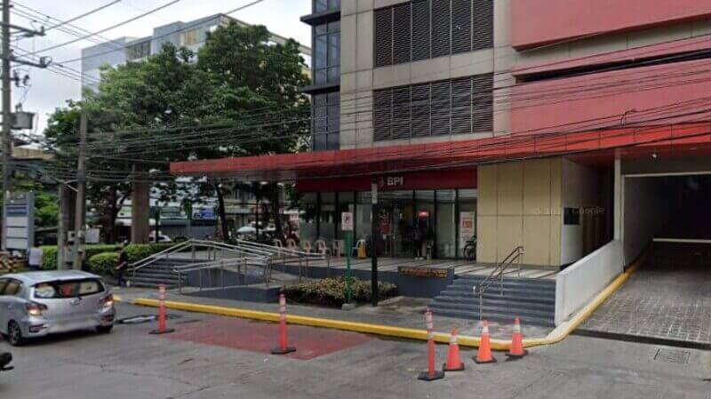 Street-level view of a BPI bank branch located on the ground floor of the Vertex One building in San Lazaro. The image shows the red-branded storefront, an accessible entrance with a ramp and stairs, and a driveway to the right. A silver car and orange traffic cones are visible on the street in the foreground