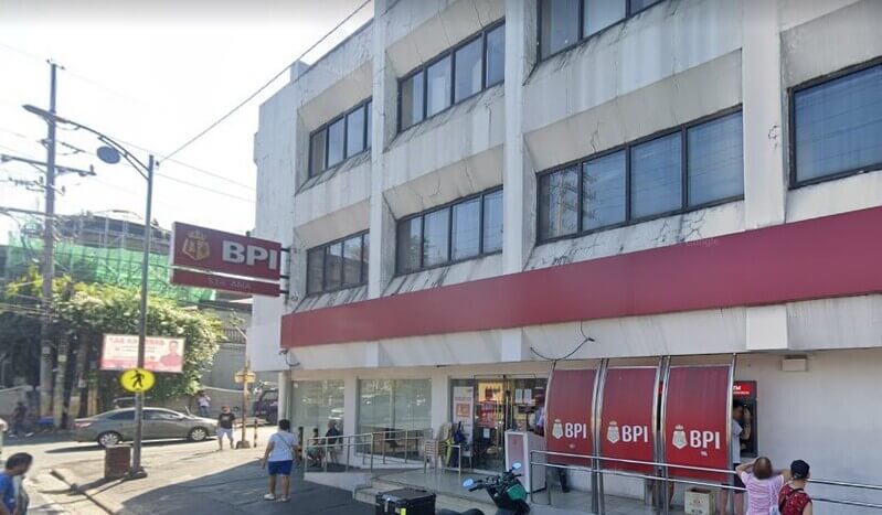 Exterior view of the BPI Sta. Ana branch featuring its red and white logo, a row of outdoor ATMs, and a white multi-story building on a sunny street corner