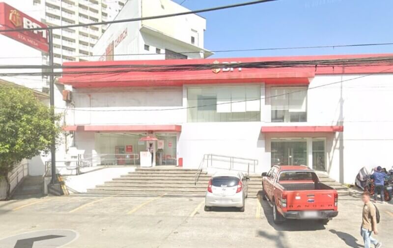 Street view of a white, one-story BPI branch in Sta. Mesa, featuring red accents, a wide concrete staircase, and parked vehicles in front under a clear sky