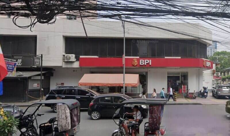 A street-level view of a BPI Vito Cruz St. Scholastica branch located on a corner, featuring its signature red signage and logo. The scene includes parked cars, local tricycles in the foreground, and pedestrians walking near the entrance under a tangled network of overhead power lines