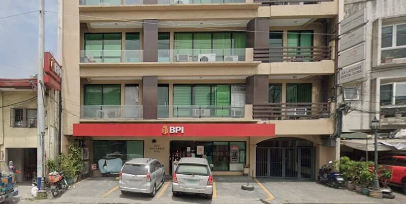 Exterior view of a multi-story tan building featuring a BPI bank branch on the ground floor with a prominent red sign. Two silver cars are parked in front of the entrance, and upper floors have balconies with green tinted windows and air conditioning units