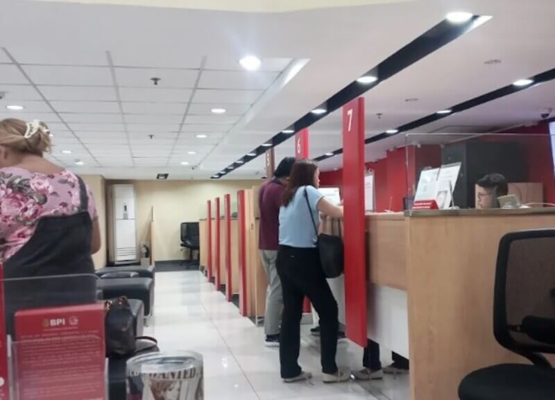 Interior view of a BPI bank branch showing customers standing at numbered service counters with red dividers under bright overhead lighting